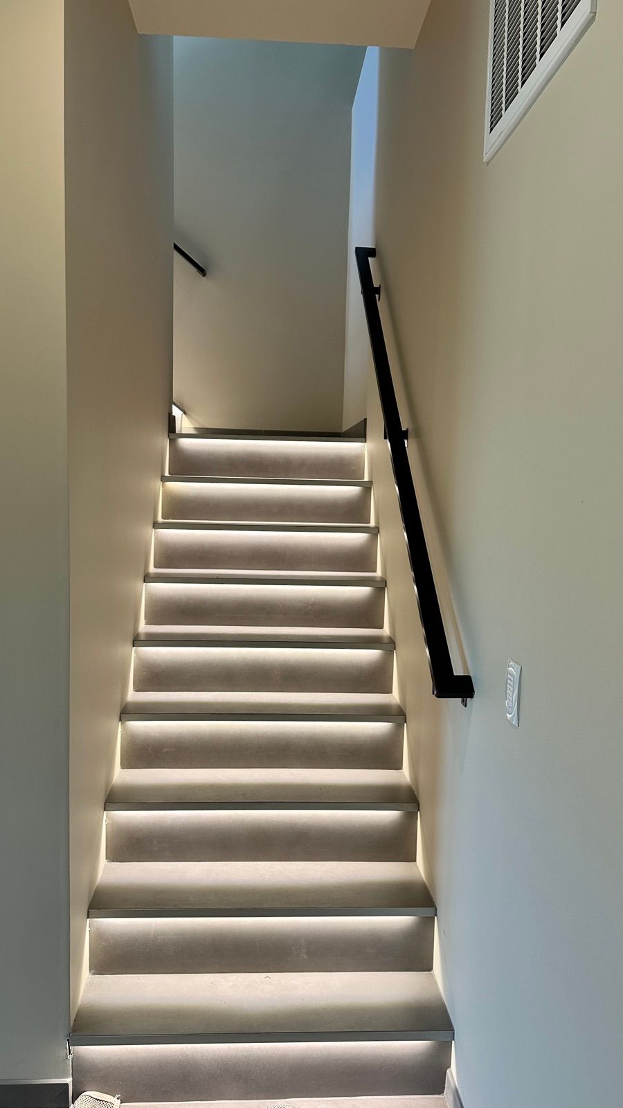 Under-tread LED step lighting on a microcement staircase with a black metal handrail.