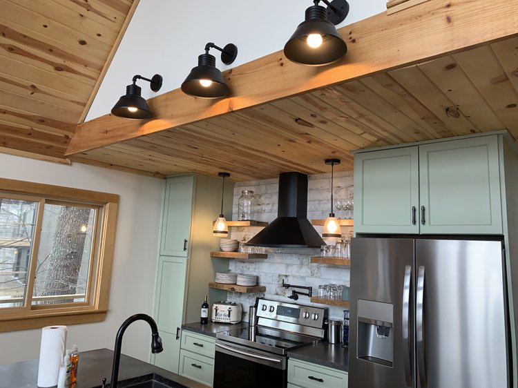 Three black gooseneck pendant lights mounted to a wood beam over an Asheville kitchen with sage cabinets and floating shelves