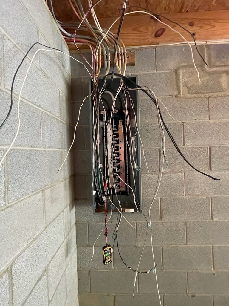 Open electrical panel in an Asheville basement with the deadfront removed, branch circuit conductors being landed on the breakers, and a meter hanging in front of the panel during diagnosis
